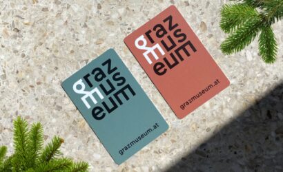 Zwei unterschiedlich farbige Karten mit dem Schriftzug „grazmuseum“ in einem gestapelten, modernen Layout. Die linke Karte ist blaugrau mit schwarzer und weißer Schrift, die rechte Karte ist terrakottafarben mit ebenfalls schwarzer und weißer Schrift. Beide Karten zeigen unten die URL „grazmuseum.at“. Die Karten liegen auf einer terrazzoähnlichen Steinfläche, eingerahmt von grünen Tannenzweigen.
