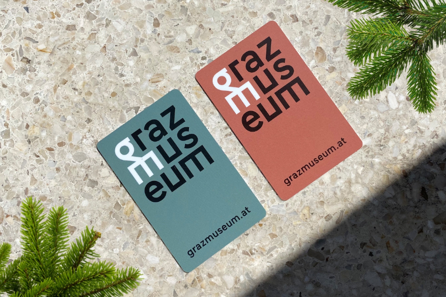 Zwei unterschiedlich farbige Karten mit dem Schriftzug „grazmuseum“ in einem gestapelten, modernen Layout. Die linke Karte ist blaugrau mit schwarzer und weißer Schrift, die rechte Karte ist terrakottafarben mit ebenfalls schwarzer und weißer Schrift. Beide Karten zeigen unten die URL „grazmuseum.at“. Die Karten liegen auf einer terrazzoähnlichen Steinfläche, eingerahmt von grünen Tannenzweigen.