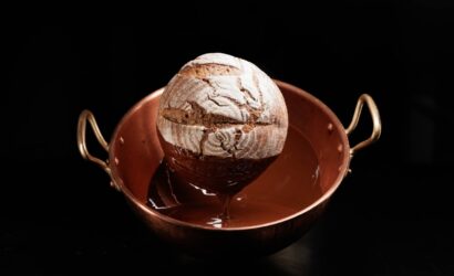 Rundes, rustikales Brot mit mehliger Kruste wird zur Hälfte in flüssige Schokolade getaucht. Es befindet sich in einem glänzenden Kupfertopf mit zwei Griffen, vor schwarzem Hintergrund. Die Szene wirkt kunstvoll und kontrastreich – eine ungewöhnliche Kombination von Brot und Schokolade.